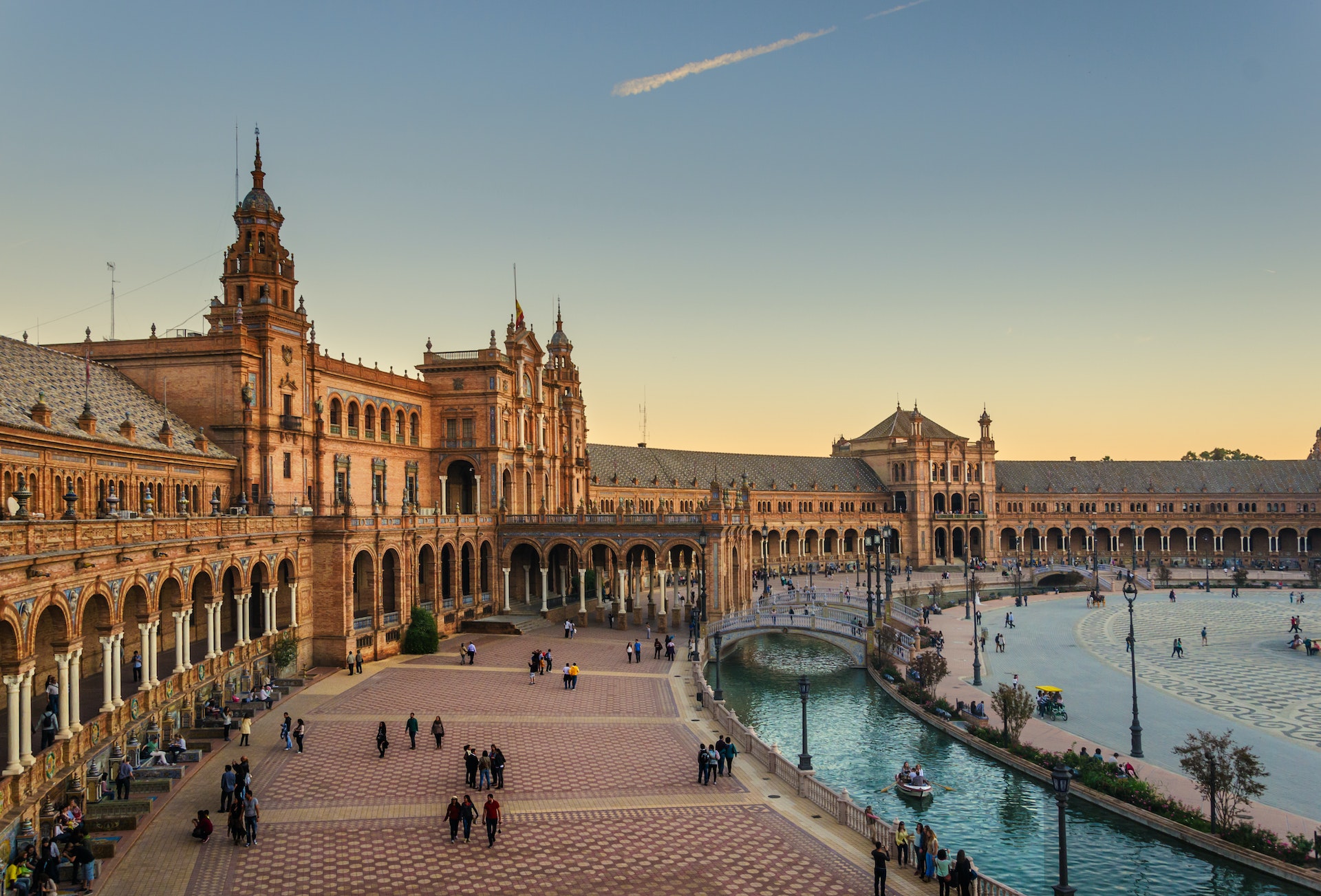 People wander around a vast square that is ringed by semicircle of buildings in a Gothic style. A small boating stream runs through the centre