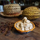 A pile of cacao fruit pods sit in a bowl, next to the bowl are roasted cacao beans. In the background is a full cacao fruit and a straw hat. 