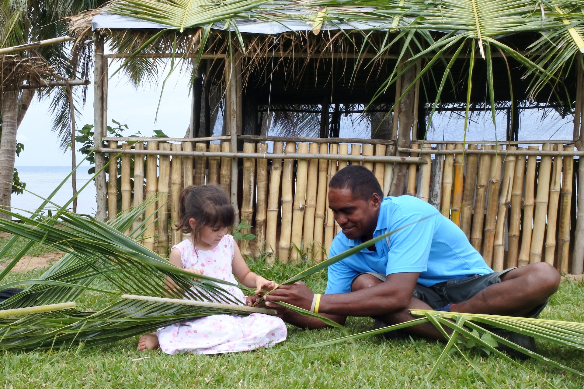 A Fijian man shows a young tourist girl how to create a basket from weaving together palm fronds