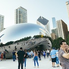 This is a horizontal color photograph of people crowded around the landmark downtown Chicago's Cloudgate in Millennial Park. The urban cityscape is reflected in the mirrored surface. A couple takes a selfie in the foreground.